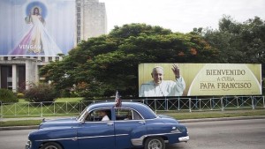 An old vehicle passes a travel ad Pope Francisco and a picture of Jesus in Havana.